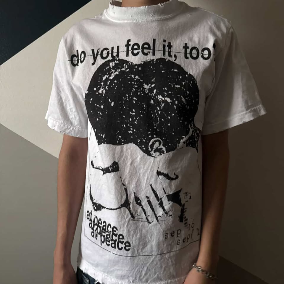 White t-shirt with black graphic and text worn by a person against a neutral background